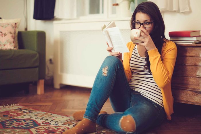Woman sitting on the living room floor reading a book and drinking from a mug.