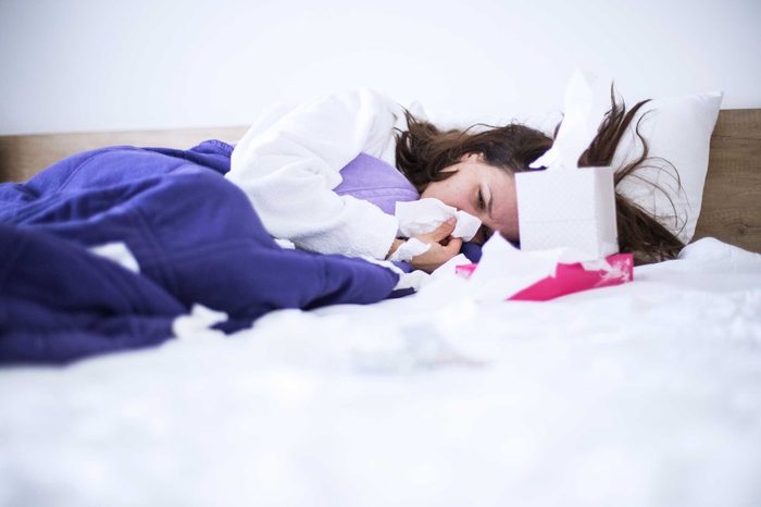 Woman in bed with a box of tissues, blowing her nose.