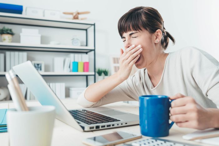 woman with a cup of coffee sitting at a desk in front of a laptop, yawning