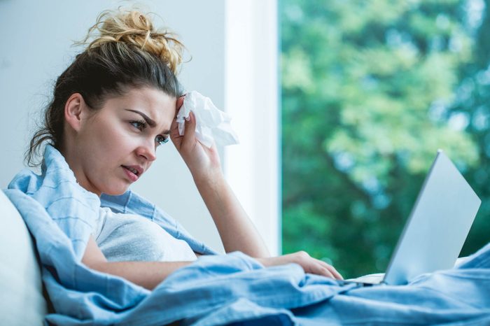 women in bed, grimacing, holding a tissue and looking at a laptop