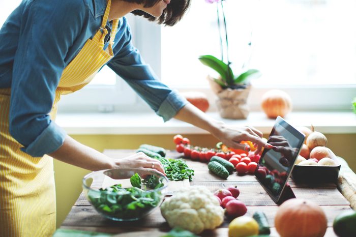 woman with apron cooking with veggies on a wooden counter, checking an iPad