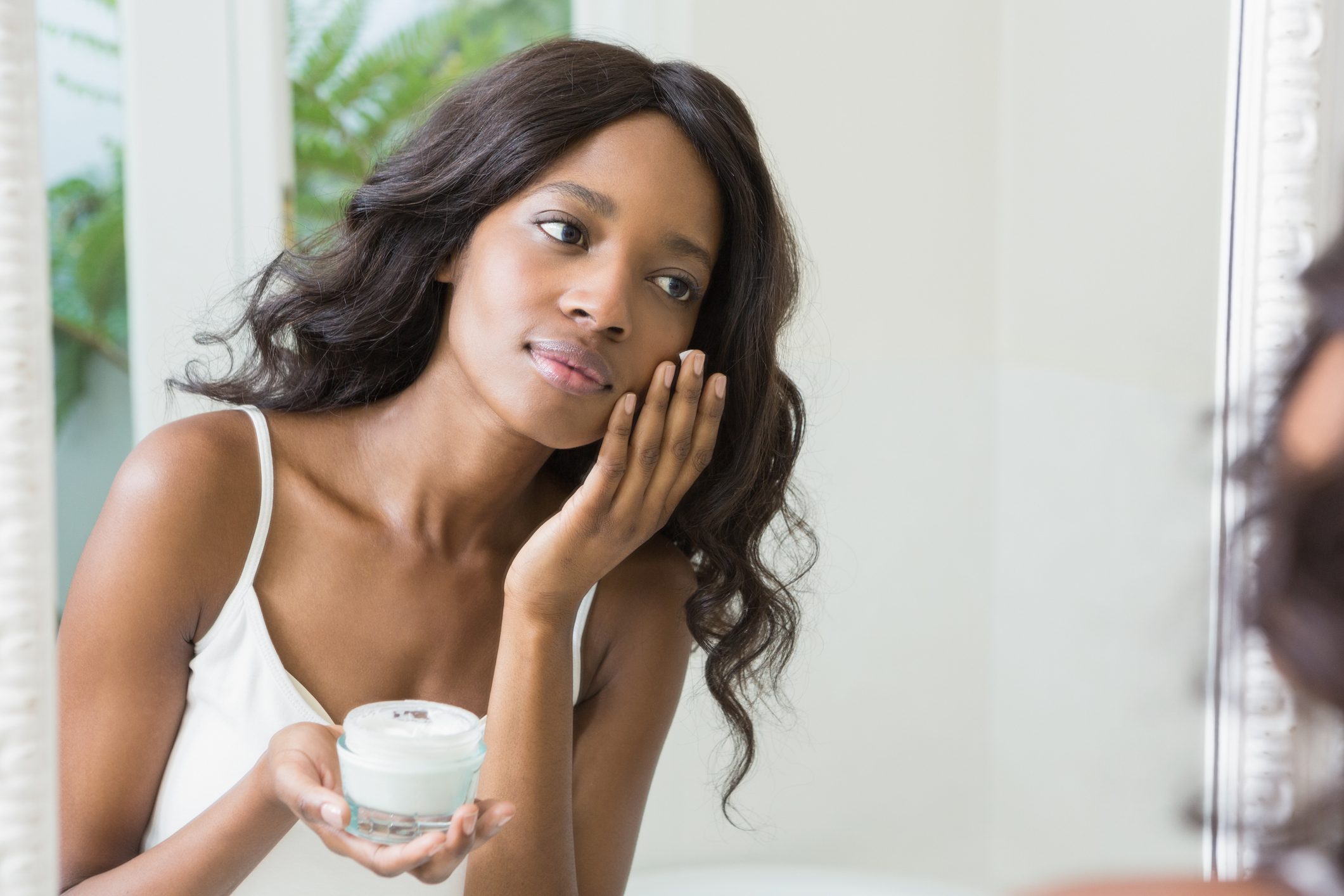 young woman looking in mirror and applying moisturizer to face