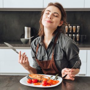 Image of pretty young woman sitting in kitchen while eating fish and tomatoes.