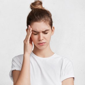 Portrait of tired exhausted young female has terrible headache after work, comes home in bad mood, being fed up with everything, needs rest, closes eyes and keeps hands on temples as feels pain