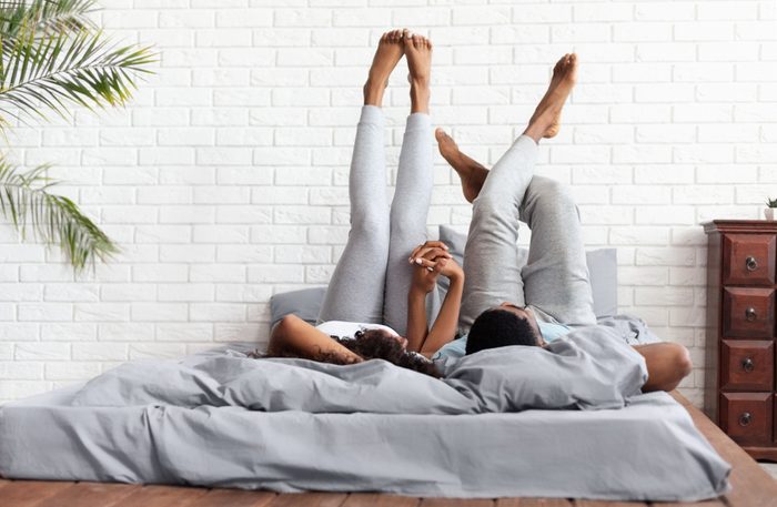 Couple in love. Young happy african-american man and woman lying on bed at home raising legs up, lazy family resting on weekend, copy space