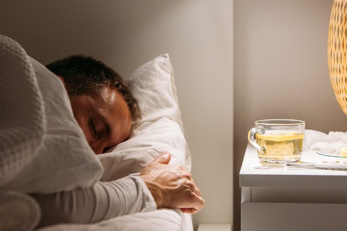 Sick man sleeps covered with a blanket lying in bed with high fever, resting at living room. He is exhausted and suffering from flu with cup of tea with lemon, medicaments. Influenza season.