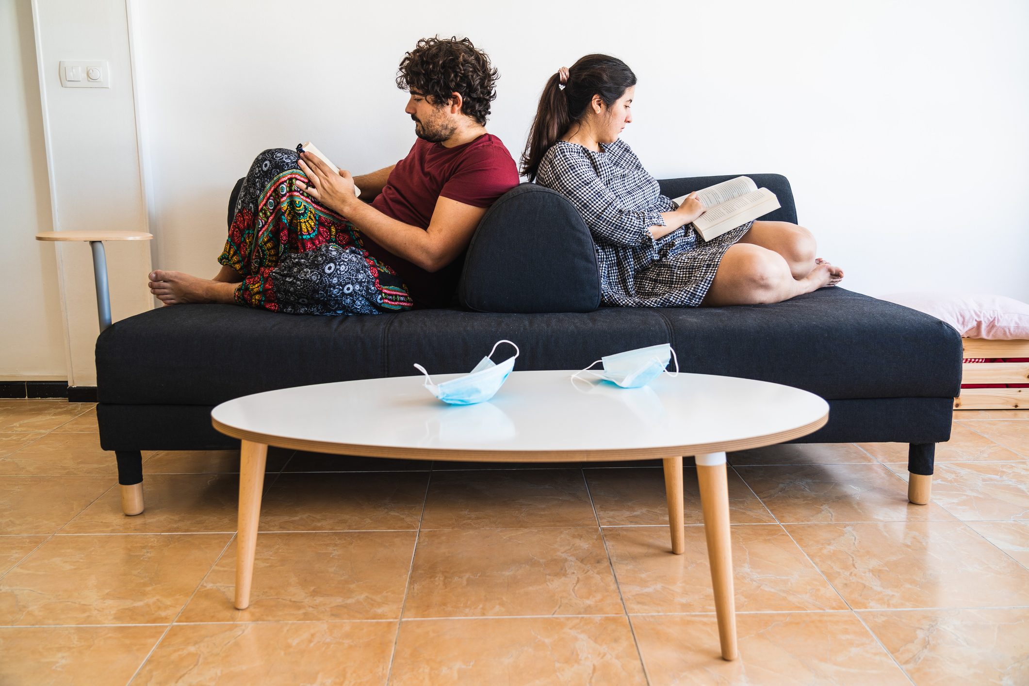 New normal situation after the 2020 coronavirus covid 19 sanitary crisis, a spanish couple rest on a black couch reading books while their surgical masks rest at the living room table