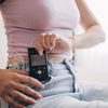 A woman adjusts the controls of her insulin pump device clipped to her jeans while sitting on a comfortable sofa in an airy and bright interior space.