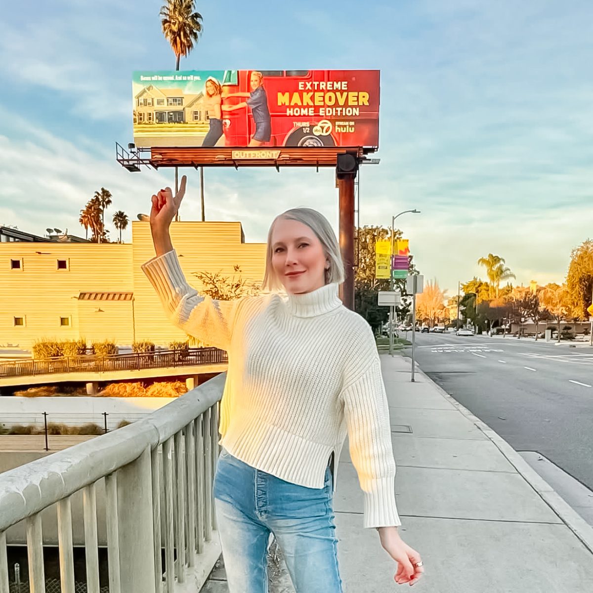 A person with light hair wearing a white sweater and blue jeans poses outdoors on a sidewalk. They are smiling and raising an arm. In the background, a billboard and palm trees are visible under a clear sky.