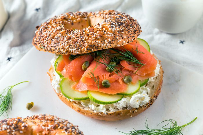 An everything bagel sandwich with cream cheese, smoked salmon, cucumber slices, capers, and fresh dill on a white surface with herb garnish nearby.